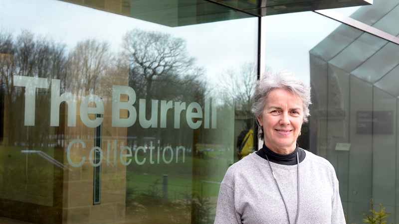 A person standing outside the Burrell collection. They have grey hair and a grey jumper with a lanyard around their neck. They are smiling at the camera.