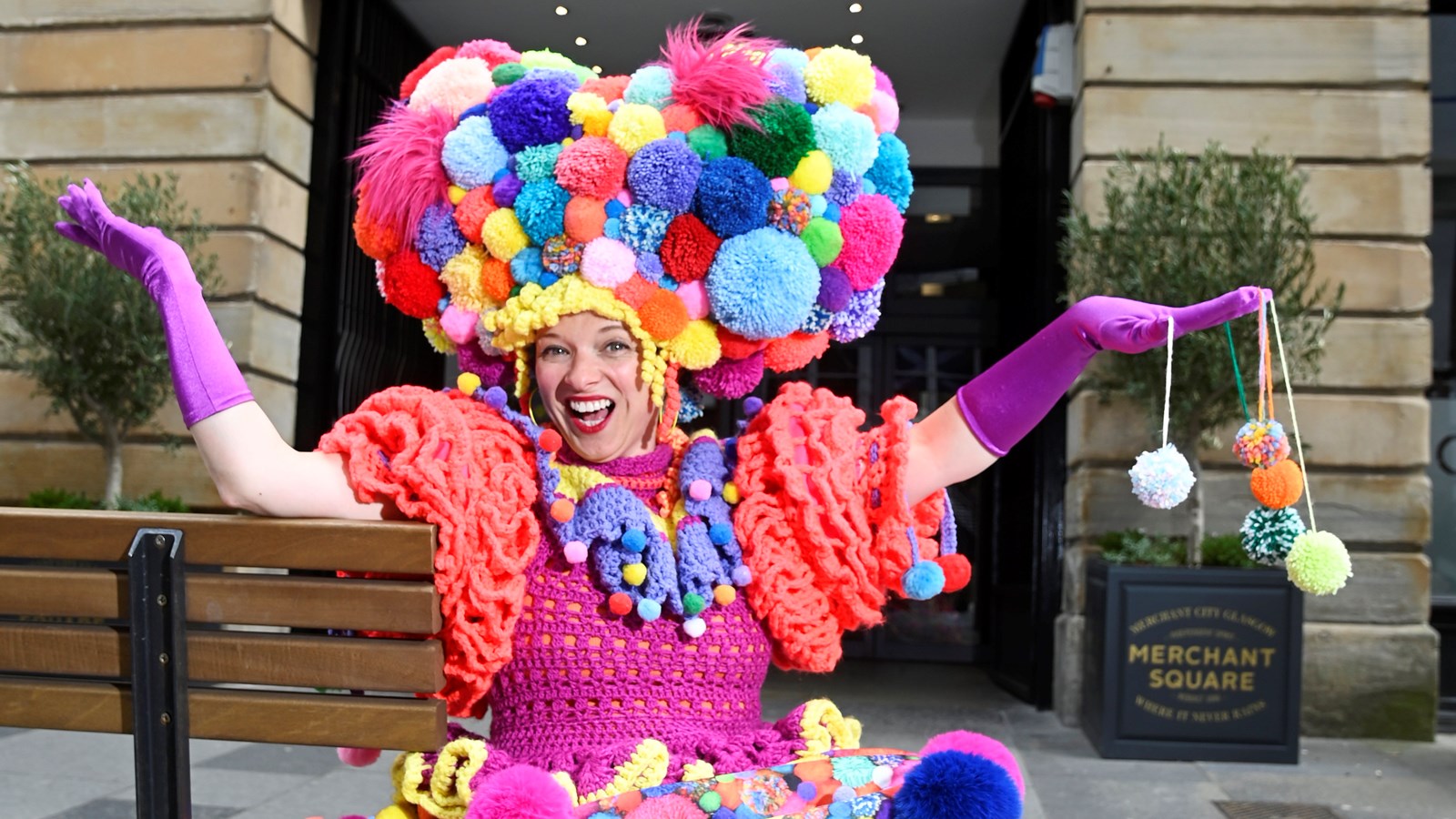 A performer wearing an outfit made of brightly coloured pom-poms sits on a bench in front of a sign which reads 'Merchant Square'