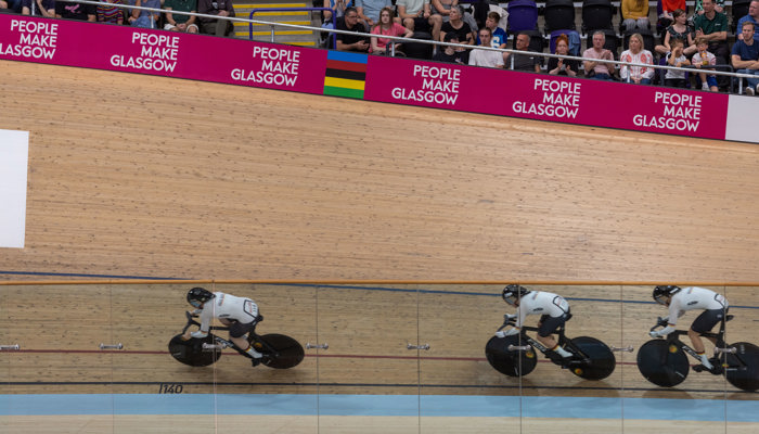 Three cyclists racing in the Sir Chris Hoy Velodrome during the 2023 UCI Cycling World Championships in front of a pink People Make Glasgow advertising hoarding.