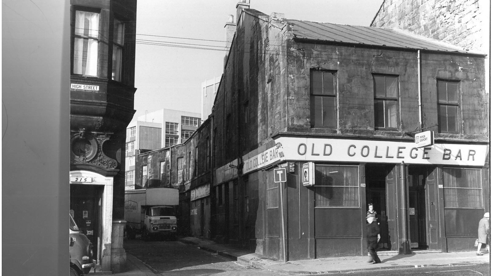 A black and white photograph of a pub in Glasgow called Old College Bar which is situated on a main road with a lane on it's right hand side. There is a parked white lorry in the lane and people walking on the pavement outside the pub.