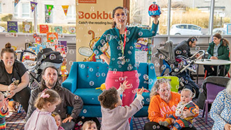 A person is holding a bubble blowing machine blowing bubbles over children who are smiling and reaching up to pop them