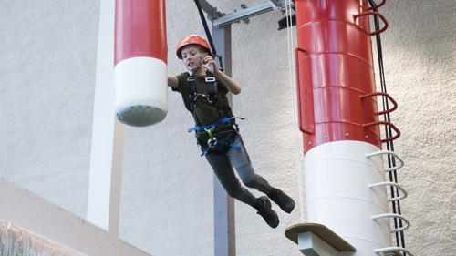 young person wearing a helmet and safety cord attached to a harness jumping through the air from a high platfrom