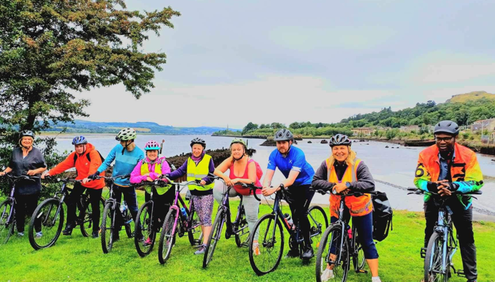 A group of adults, who are a mix of genders and ethnicities, from Drumchapel Cycle Hub on their bikes in front of a large body of water and greenery.