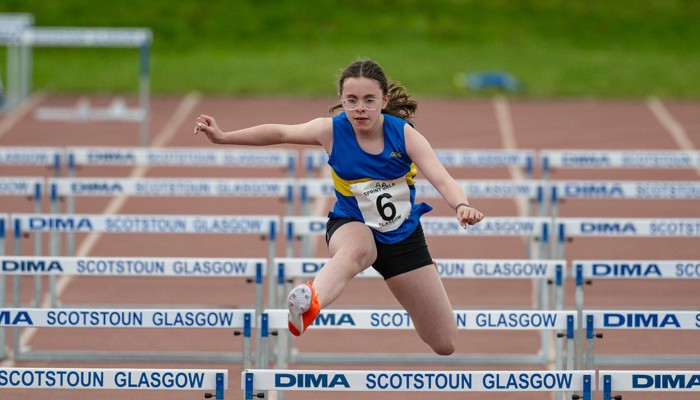 A young Shettleston Harrier athlete jumping over hurdles during a race.