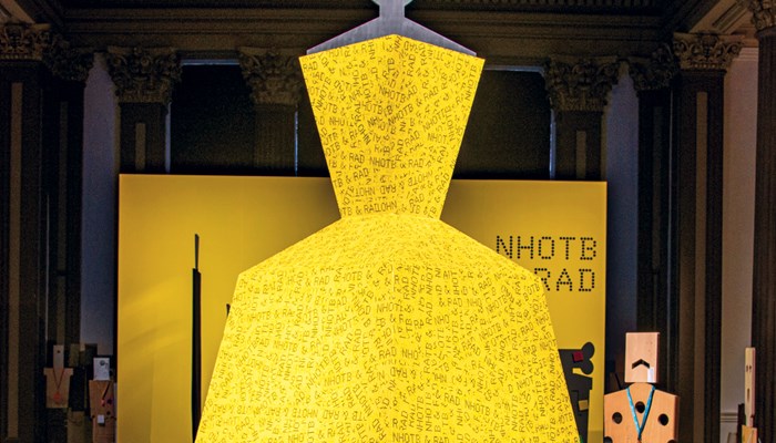A bright yellow ballroom type dress on a black plinth during the installation of a Beagles and Ramsay exhibition at Glasgow Gallery of Modern Art, 2023. The name of the exhibition, NHOTB & RAD, can be seen in the background..