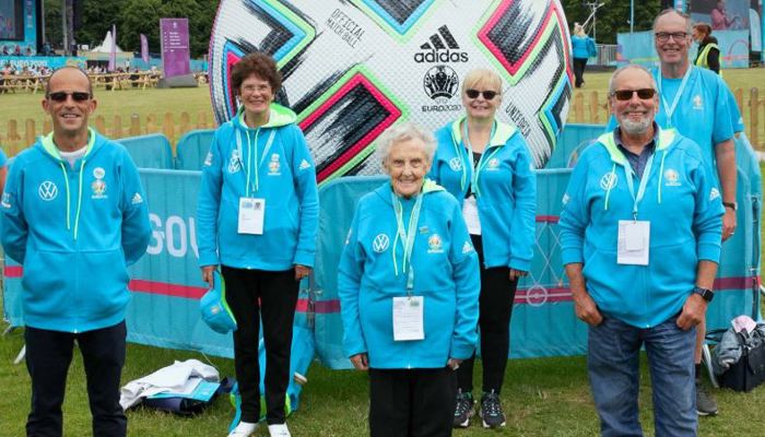 Six volunteers, who represent different ages and genders, standing in front of a giant Adidas football in the Glasgow Green fan zone for Euro 2020.