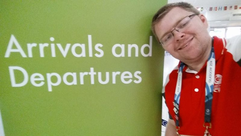 Andrew McVie wearing his red Clyde-sider uniform in front of the arrivals and departures sign at Glasgow Airport during the 2014 Commonwealth Games.