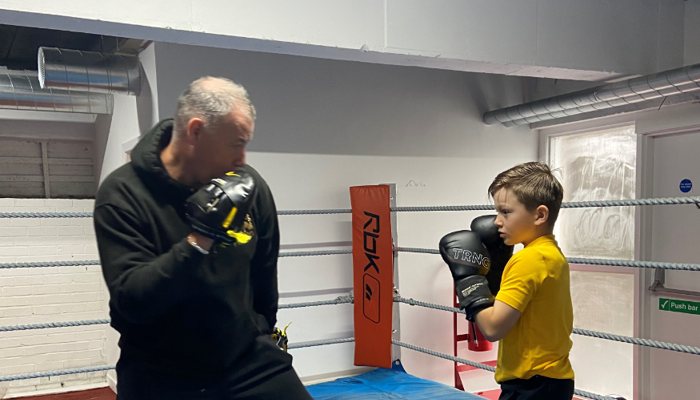 An adult teaching a child boxing, they are both wearing black boxing gloves and standing in the middle of a boxing ring.