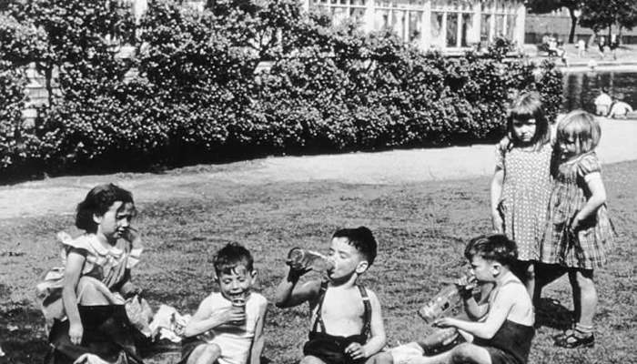 Black and white photo showing 6 children in a park having a picnic, three of the children are boys and are drinking from bottles of juice.