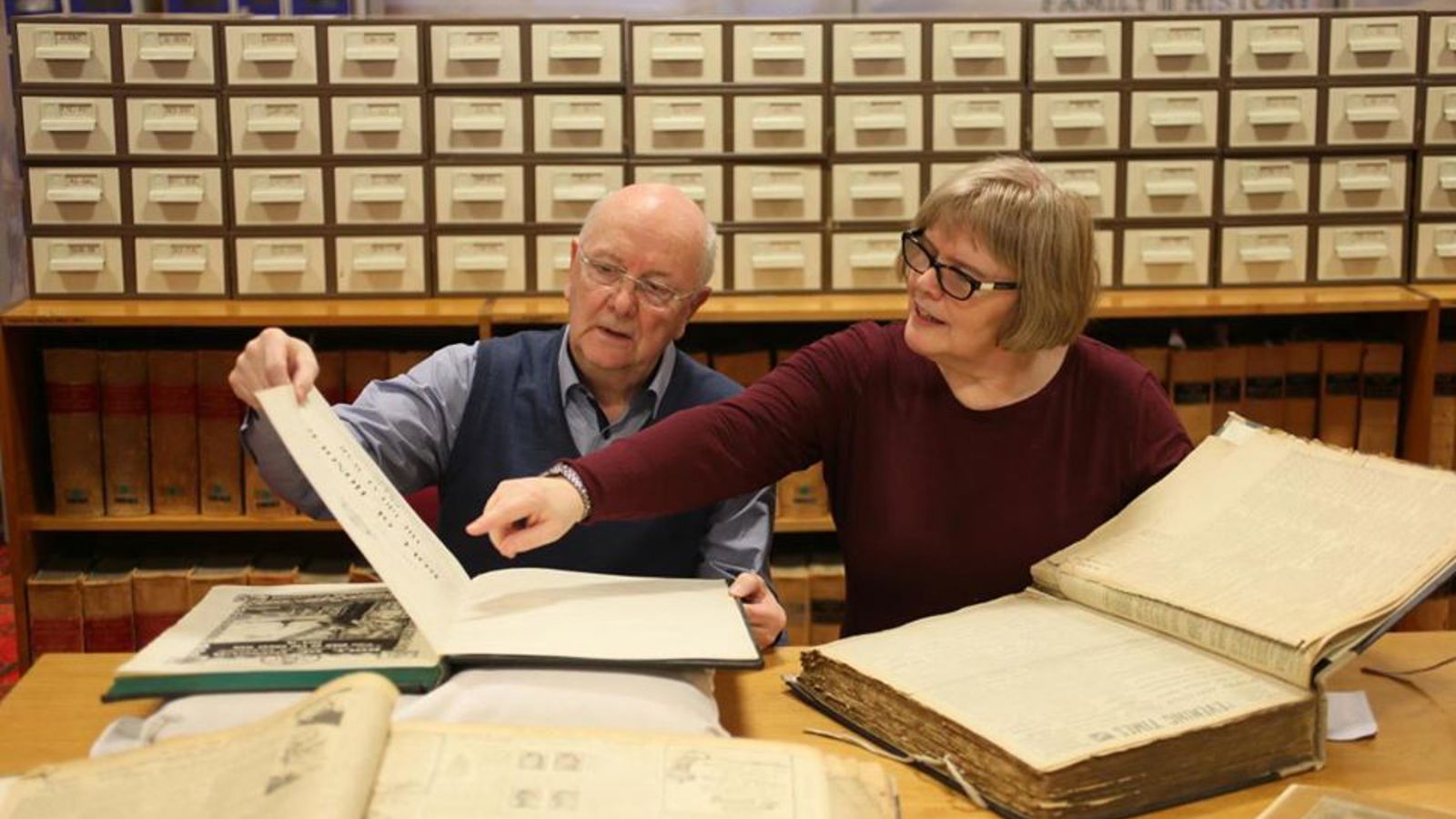 Two people sitting at a desk with two large old books in front of them with filing cabinets behind them in the background