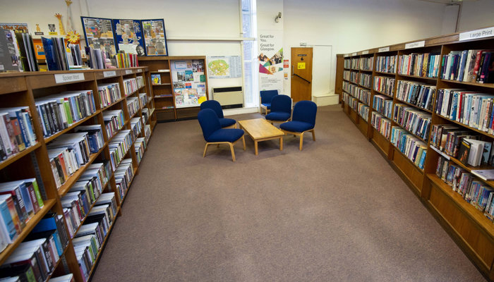 Two rows of wooden bookshelves at each side of the picture. In the middle is a coffee table with 4 blue soft chairs