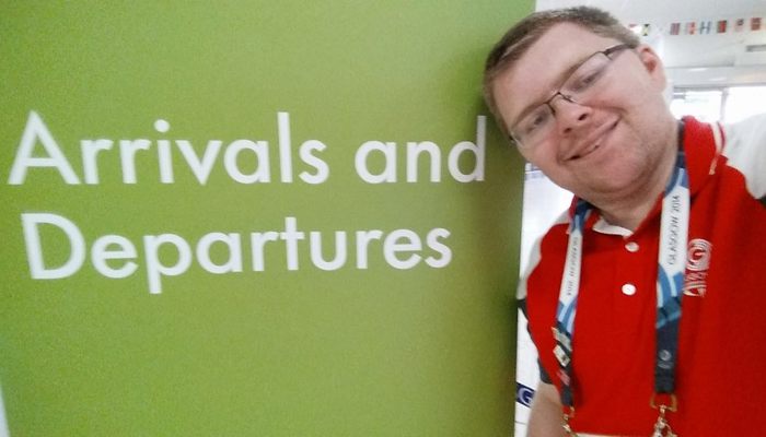 Andrew McVie wearing his red Clyde-sider uniform in front of the arrivals and departures sign at Glasgow Airport during the 2014 Commonwealth Games.