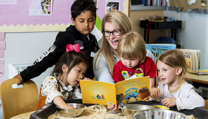 Adult sits at a table reading a book with four children around her