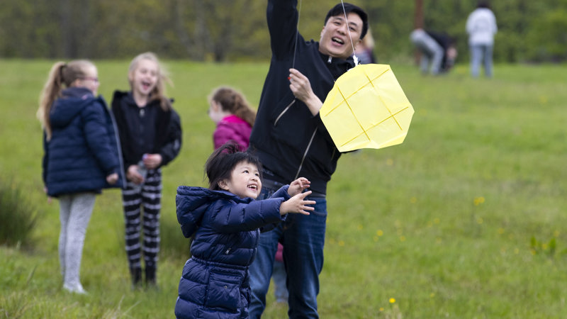 A child and adult laugh together as they play with a kite. Behind them, in a grassy setting, more children laugh together