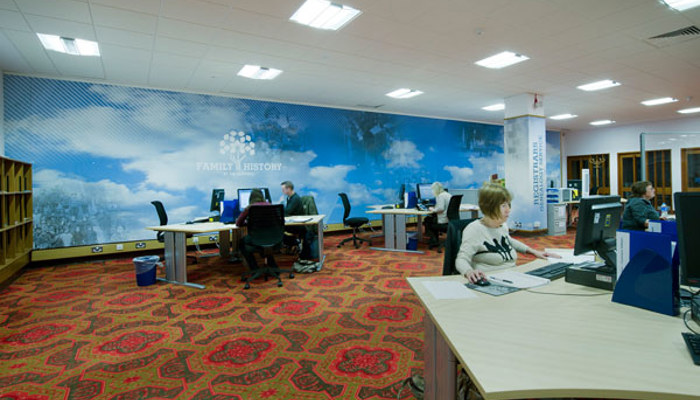 A room in the Family History centre at The Mitchell Library featuring multiple desks with PCs and bookcases, there are a few people sitting at the desks looking at the computer screens.