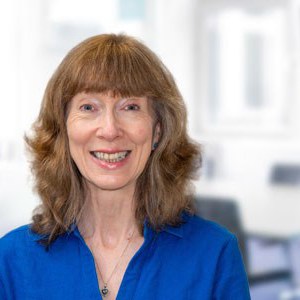 Lynn Bradley is wearing a blue top and is smiling at the camera, she is in front of a bright window