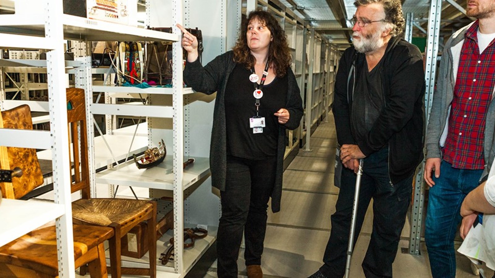 A selection of people looking at some museums collections in a storeroom. They are looking at two chairs and the guide is pointing at the exhibits and speaking to the tour group