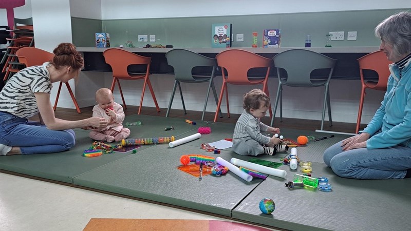 Two adults with two small babies sitting on mats on the floor with lots of balls and other items scattered around on the floor, a row of chairs are lined up against a wall in the background