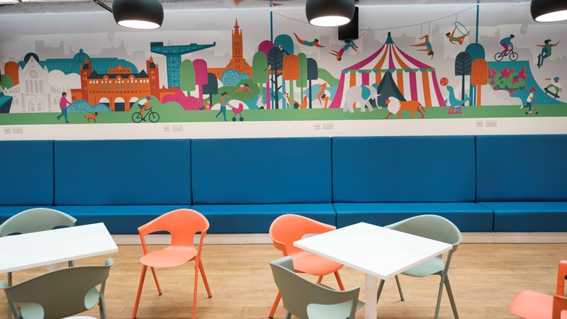 Tables with bright orange and green chairs with a colourful circus themed murals on the back wall