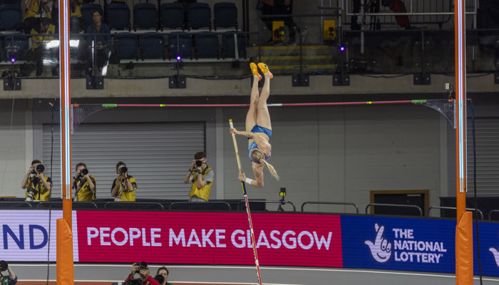 A pole vaulter attempting a clearance during the World Athletics Indoor Championships, behind the crash mat is a pink People Make Glasgow advertising hoarding.