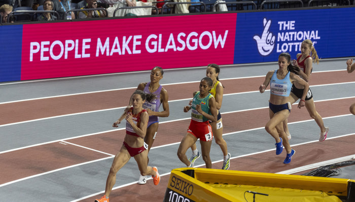 A group of athletes racing during the World Athletics Indoor Championships in Glasgow, they are in front of a pink People Make Glasgow advertisting hoarding.