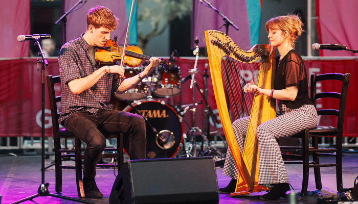 Two musicians play the violin and the harp in front of signs which read 'Glasgow: UNESCO City of Music'