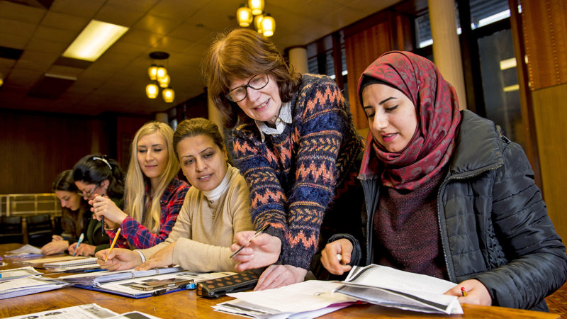 group of women of different nationalities sitting around a desk reading and writing with one women leaning over to check their work