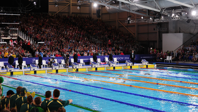 Swimmers racing at the Tollcross International Swimming Centre during the Glasgow 2014 Commonwealth Games. There is a large crowd sitting and standing in the stands watching the competition unfold.