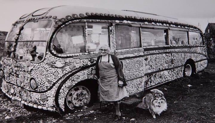 Black and white image of Flora with Shell the dog. They are standing in front of her bus that has been completely decorated in shells.