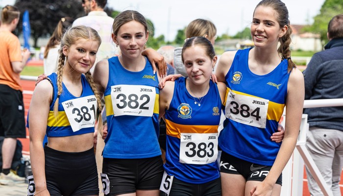 Four young Shettleston Harrier athletes after a race standing in a line with their arms round each other. They are wearing the blue and yellow club colours and racing numbers on their front.