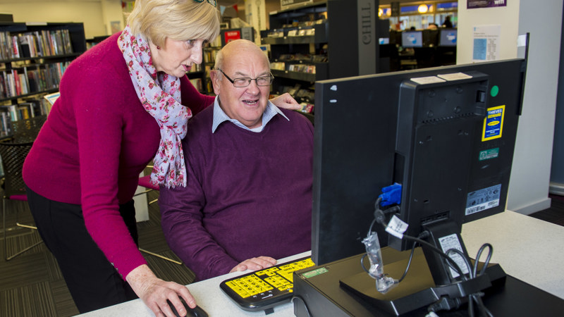 Two people in a library looking at a computer. One is sitting in front of the computer, the other is standing, leaning over and holding the computer mouse.