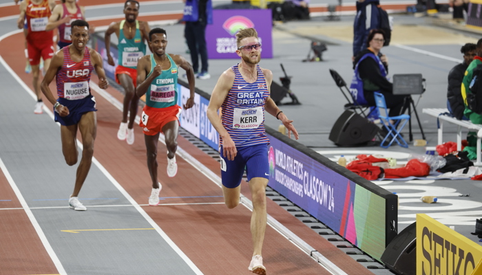 Josh running down the home straight on the track at the Emirates Arena during the 2024 World Athletics Indoor Championships. He is wearing a Great Britain vest and sunglasses, other athletes from other competing countries are behind him.