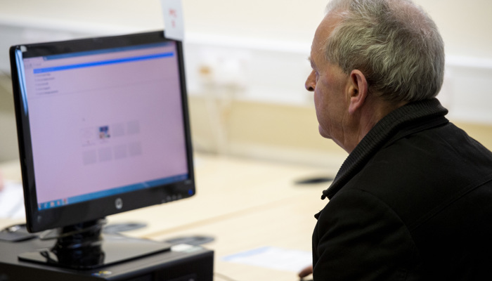 An adult sits at a PC. They have grey hair and a black jacket.