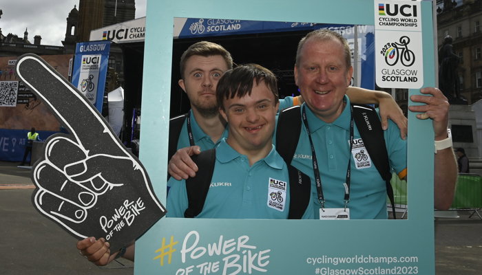 Three volunteers in Glasgow's George Square during the UCI Cycling World Championships, they are holding a picture frame and one is holding a large foam finger.